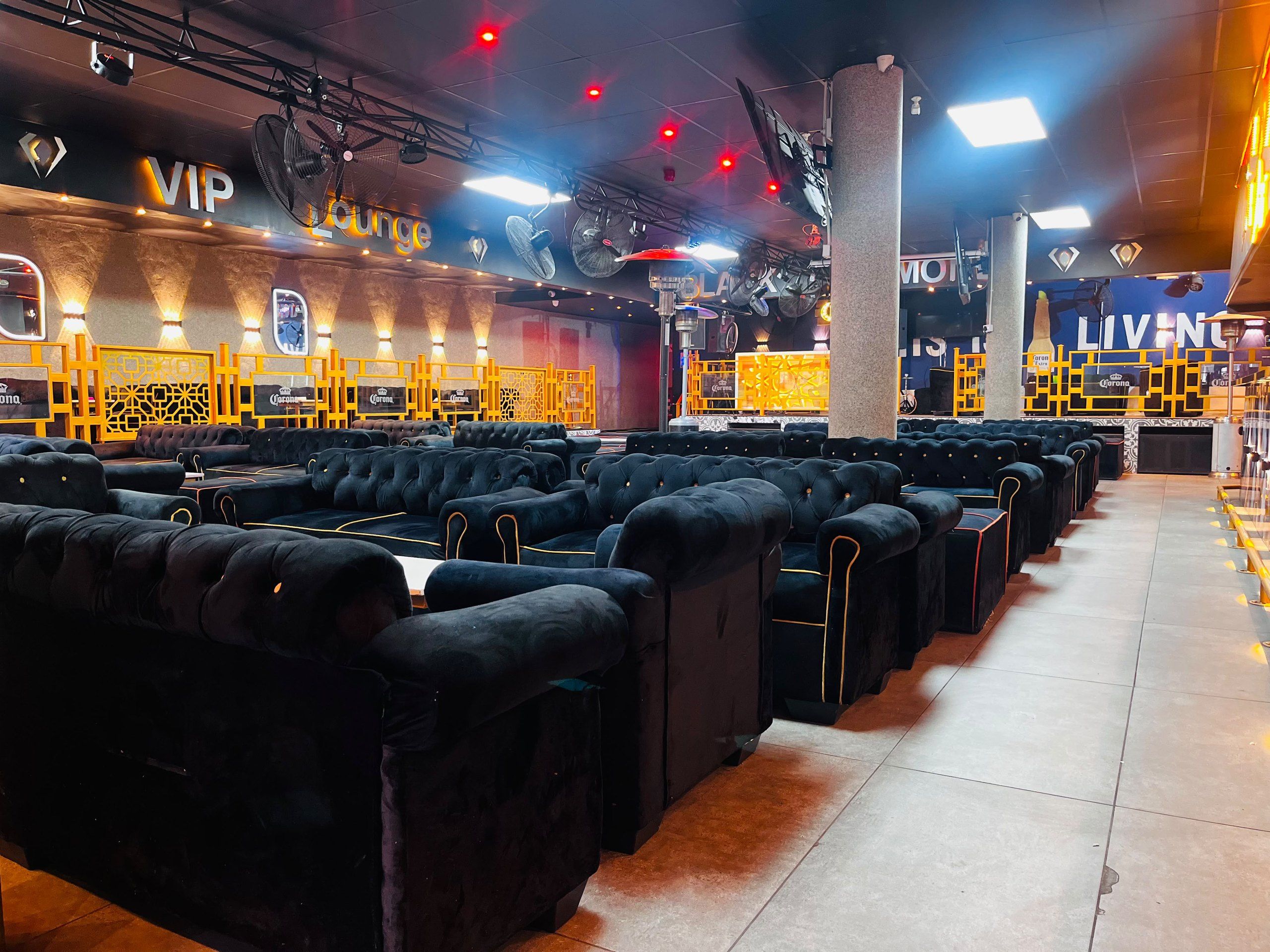 The lounge floor at Black Diamond: rows of black buttoned velvet couches receding into the room, an illuminated VIP Lounge sign in gold above a stone-clad wall, red ceiling spots overhead and a lit Corona marquee to the right.