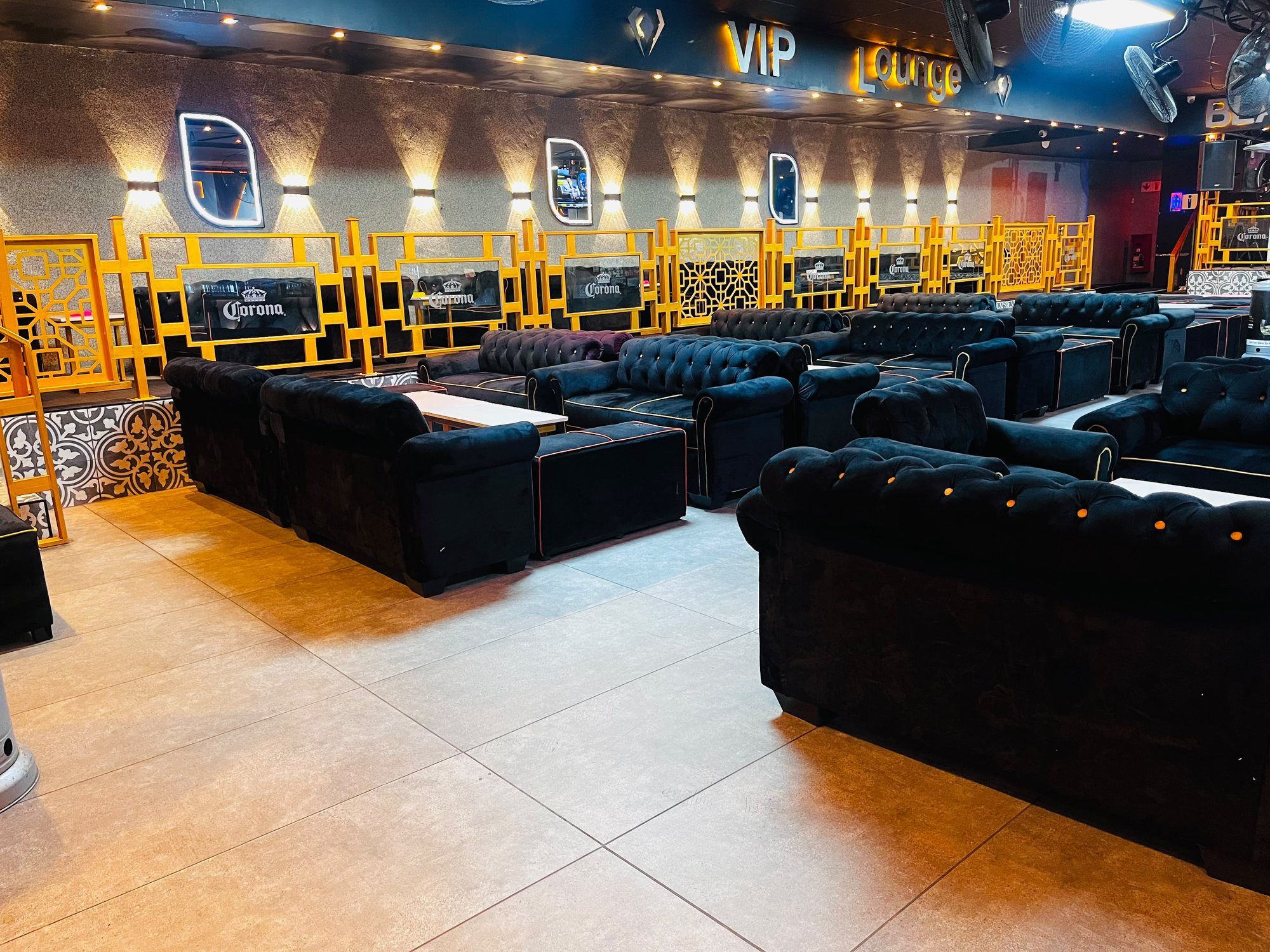 The VIP lounge: an illuminated VIP Lounge sign in gold above a stone-clad wall washed by uplights, a gold balustrade, and rows of black buttoned velvet couches on tiled flooring.